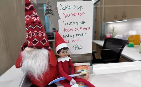 An elf on a shelf holding a toothbrush telling children, "Santa says you better brush your teeth or they'll end up black as coal!"